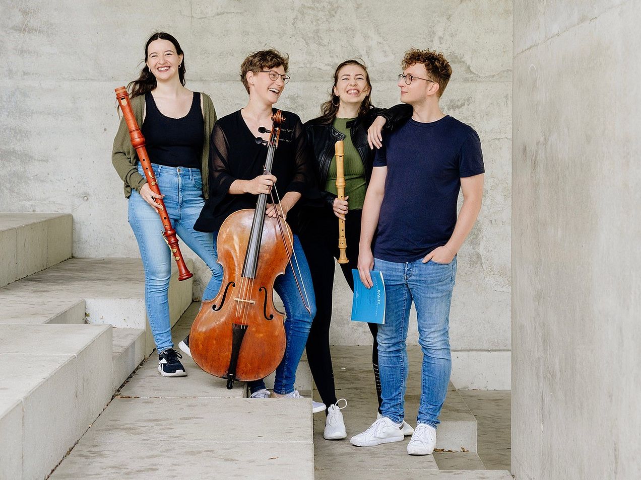Die vier Mitglieder des Ensembles (Tabea Wink und Marie Erndl (Blockflöten), Anna Maria Rudolph (Cello) und Dominik Heidl (Cembalo))  stehen nebeneinander mit ihren Instrumenten auf Beton-Stufen und lachen. 