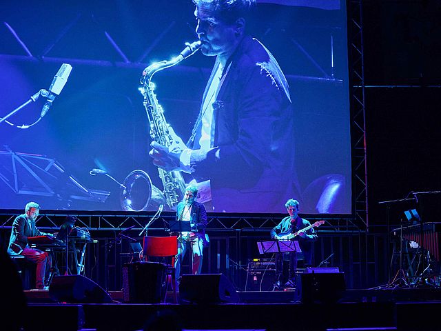 Professor Patrick Stadler steht in der Mitte, neben ihm ein Gitarrist und ein Pianist des Nikel-Ensembles. Auf der Leinwand hinter den Musikern ist ebenfalls Prof. Patrick Stadler mit seinem Saxophon zu sehen. 