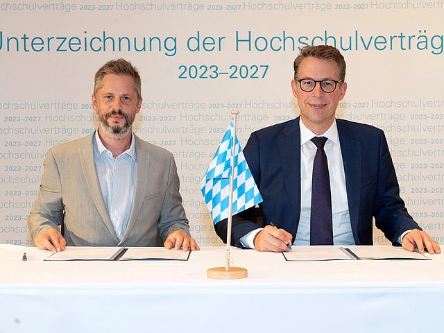 Prof. Rainer Kotzian und Markus Blume sitzen an einem Tisch. Zwischen ihnen steht eine kleine bayrische Flagge, vor ihnen liegen die unterzeichneten Verträge. 