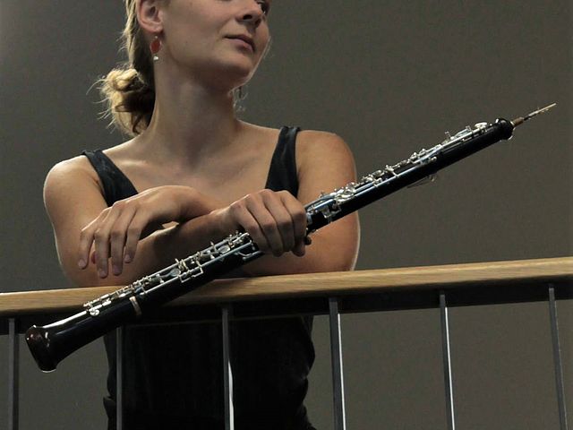 Porträt von Magdalena Steinbauer, die ihre Arme über ein Geländer legt und mit der rechten Hand ihre Oboe hält. Sie schaut freundlich lächelnd zur Seite. 