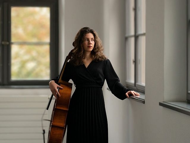 Die Cellistin im schwarzen, langen Kleid steht in einem Gang und schaut nach rechts aus dem Fenster. Einen Arm hat sie auf eine Fensterbank gelegt, mit dem anderen Arm hält sie ihr Cello fest. 